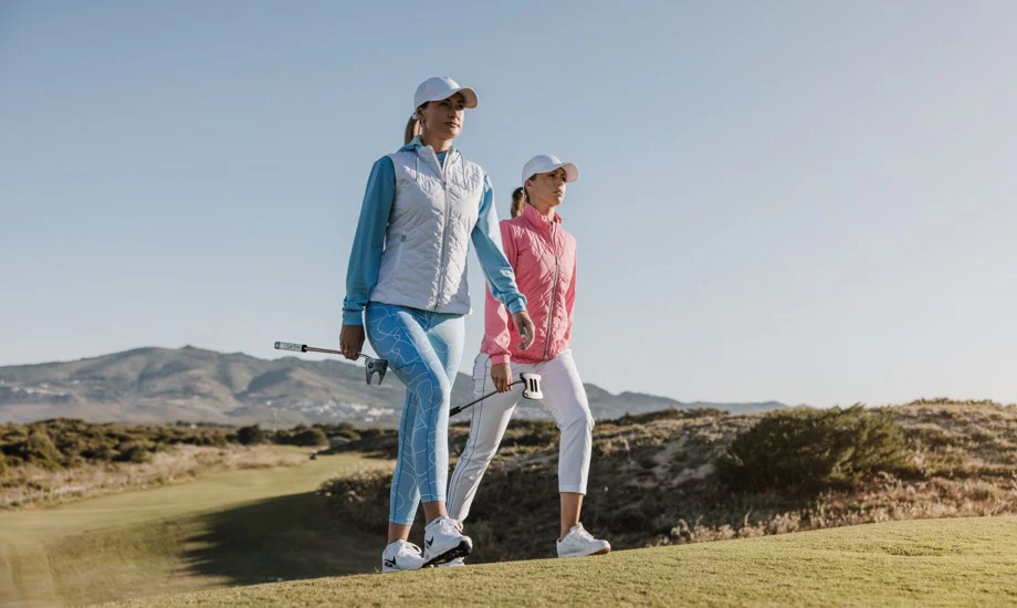 Two golfers walking a coastal fairway in layered golf apparel: a white quilted vest over a blue base layer and a pink padded jacket