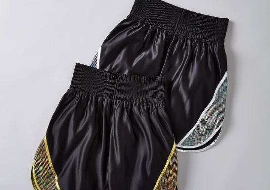 Pair of black professional boxing trunks with wide gathered waistbands, gold side piping, and contrast side panels