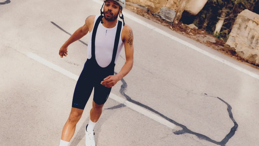 Cyclist in black bib shorts with wide white bib straps and a white base layer, standing on a road