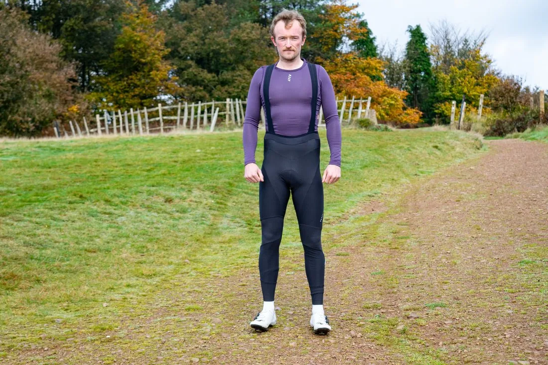 Cyclist standing outdoors in black thermal bib tights and a long-sleeve purple jersey on a cold autumn day