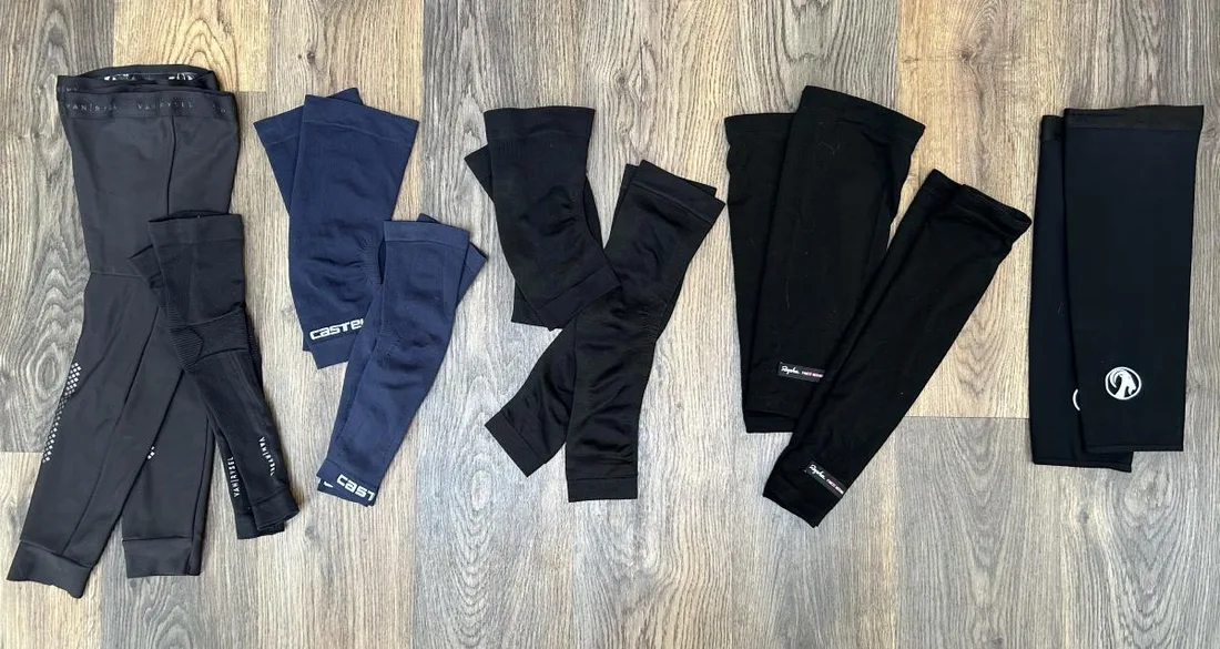Flat lay of cycling range pieces: leg warmers, navy knee warmers, black arm warmers and thermal leg covers on a wooden floor