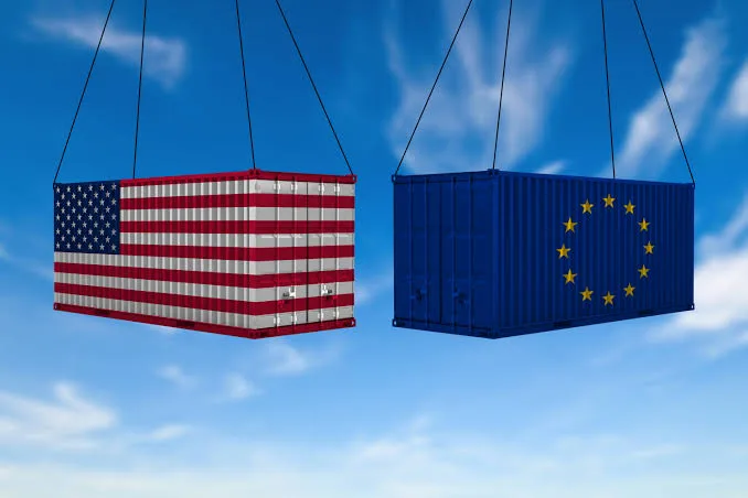 Two shipping containers hoisted by crane against a blue sky, one painted with the US flag and one with the EU flag, representing transatlantic apparel export