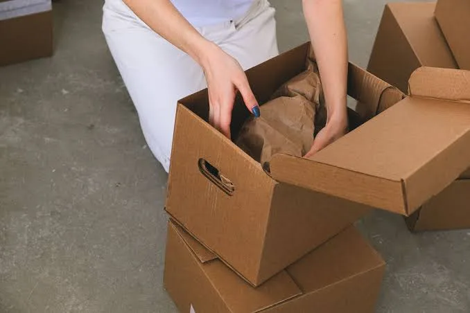 A person packing a cardboard box with crumpled kraft paper filling, surrounded by other cartons on a warehouse floor