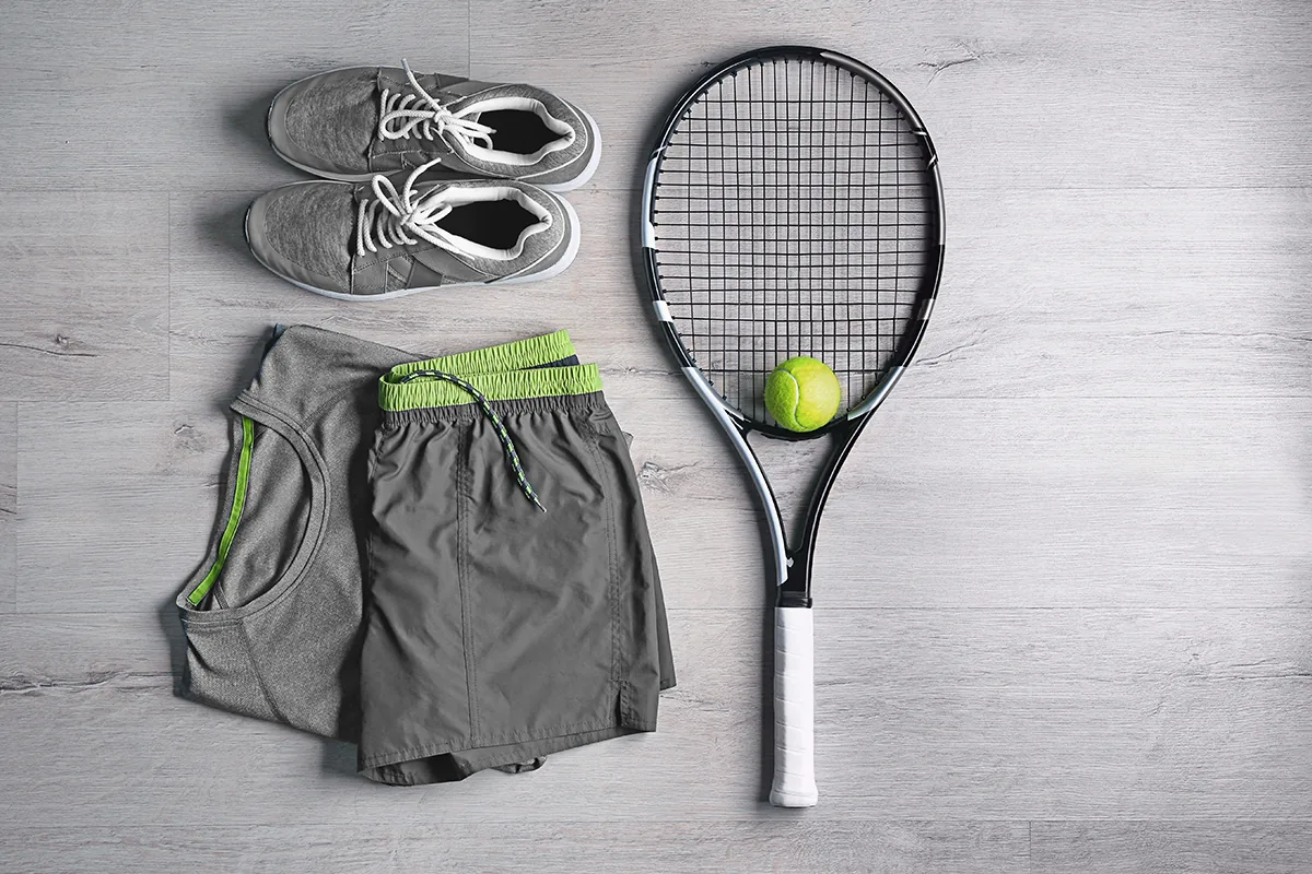 Flat lay of tennis kit: grey tank top, shorts with a lime waistband, grey sneakers, racket and tennis ball